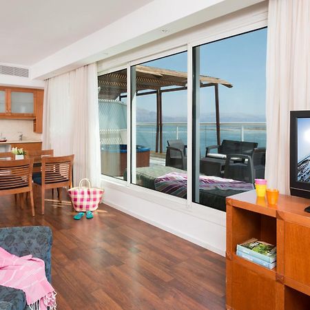 โรงแรมลีโอนาร์โด คลับ เดด ซี ออล อินคลูซีฟ เนเว โซฮาร์ ภายนอก รูปภาพ The photo depicts a modern living space with a view. On one side, there is a comfortable seating area with a couch and a television cabinet. The TV is turned on, showing a scenic image. 

In the background, there is a dining area with a wooden table 