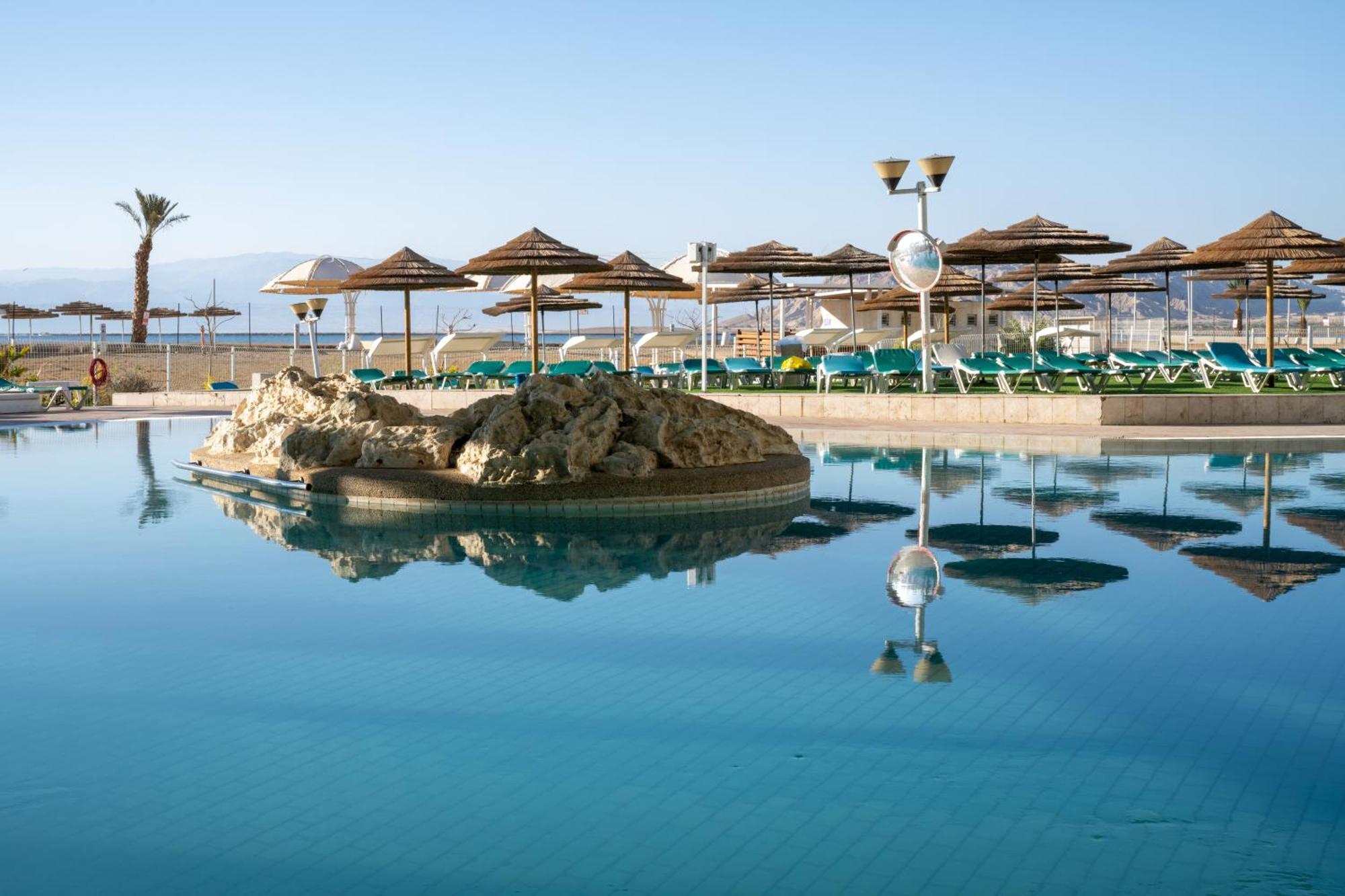 โรงแรมลีโอนาร์โด คลับ เดด ซี ออล อินคลูซีฟ เนเว โซฮาร์ ภายนอก รูปภาพ The photo shows a tranquil swimming pool area. It features a large, reflective pool with a small rock formation in the center. Surrounding the pool are several sun loungers with green cushions and a series of thatched umbrellas providing shade. In th