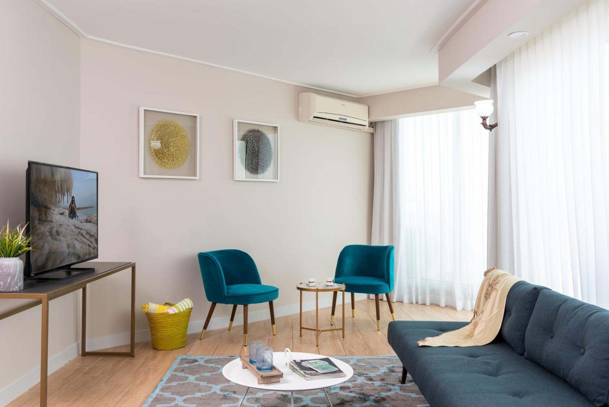 โรงแรมลีโอนาร์โด คลับ เดด ซี ออล อินคลูซีฟ เนเว โซฮาร์ ภายนอก รูปภาพ The photo depicts a modern living room setting. The room features a dark blue sofa with a light throw blanket draped over it. There are two stylish teal accent chairs positioned around a small round coffee table, which holds various items. A flat-scr